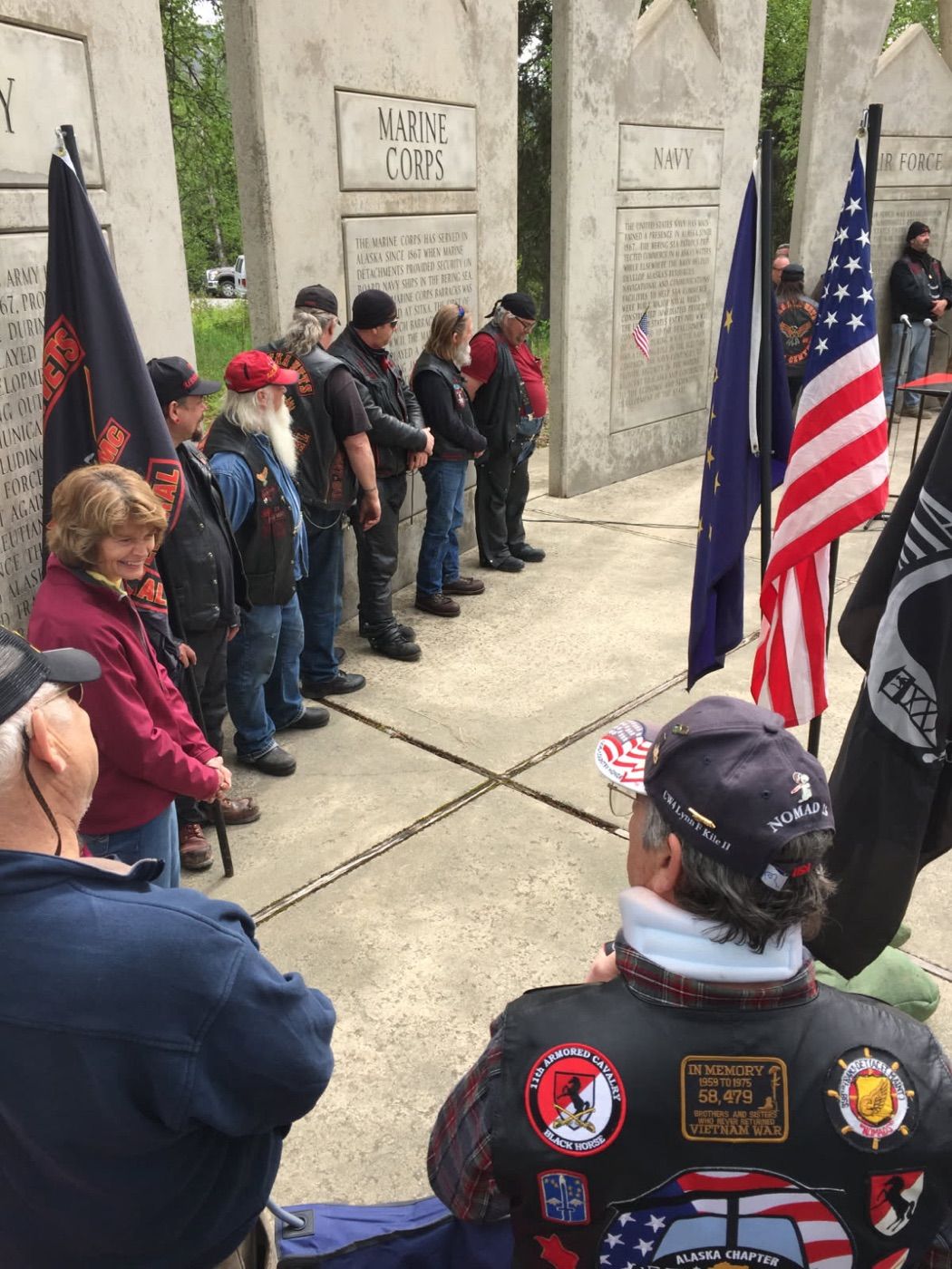 Veterans in riding vests standing at the military branch memorial walls at Byers Lake, with the Alaska and American flags