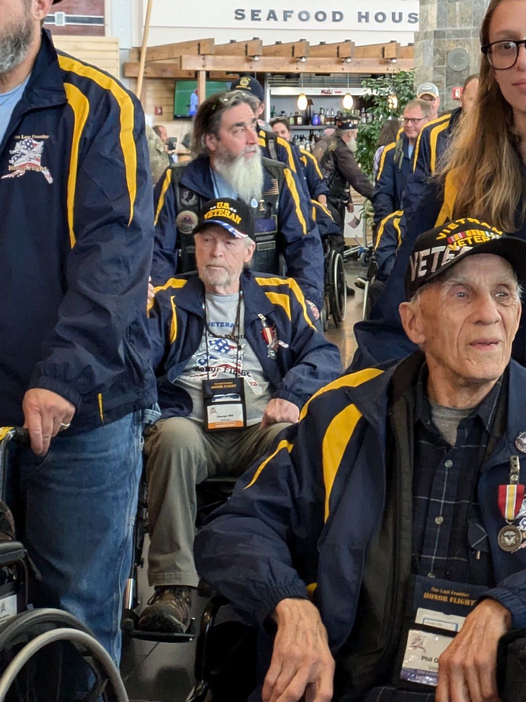 Honor Flight Veterans arriving in wheelchairs, with volunteers beside them